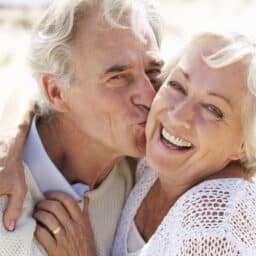 Senior Couple Walking Along Beach Together Smiling To Camera.